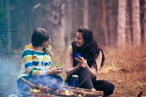 Deux femmes savourent un café près d'un feu de camp dans une forêt, profitant de la chaleur et de la convivialité de la nature.